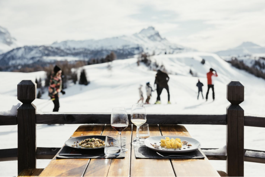 A table with plates of food and glasses of wine are waiting for dinners as people ski in the background